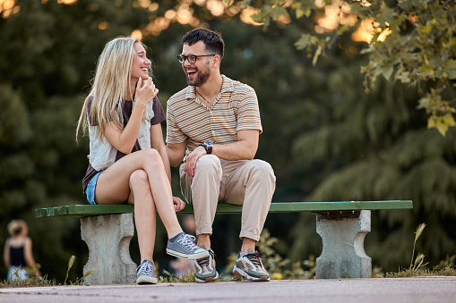 Young cheerful couple communicating while relaxing on a bench at the park.