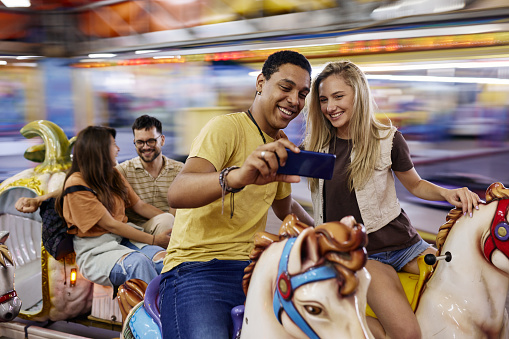 Young happy couple having fun while taking a selfie with cell phone during carousel ride at amusement park. Their friends are in the background. Blurred motion.