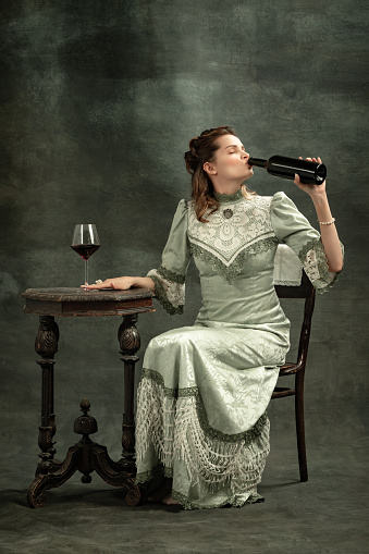 Tasting red wine. Conceptual portrait of young charming girl in image of medieval royal person or viscountess drinking wine isolated on dark background. Comparison of eras concept, fashion, art, ad.