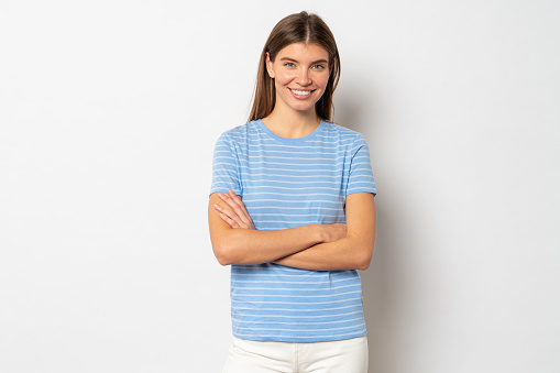 Positive lovely confident young woman with brown hair in blue striped t-shirt standing with folded hands, looking at you against white studio background. Human sincere emotions concept