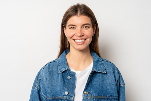 Head and shoulders portrait of happy youthful beautiful woman with wide sincere candid smile, wearing casual jean jacket standing over white background