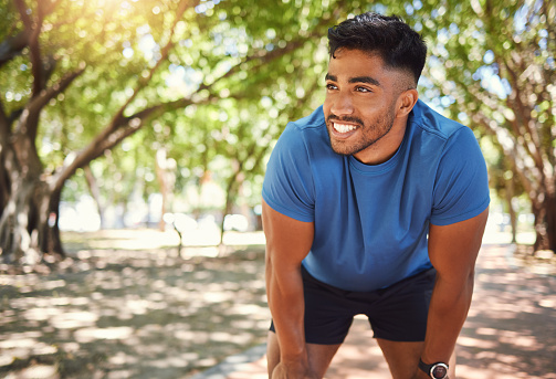 Happy athletic mixed race man smiling while standing with his hands on his knees in a park. Hispanic male resting after his run. Fit man pleased with his performance while exercising outdoors