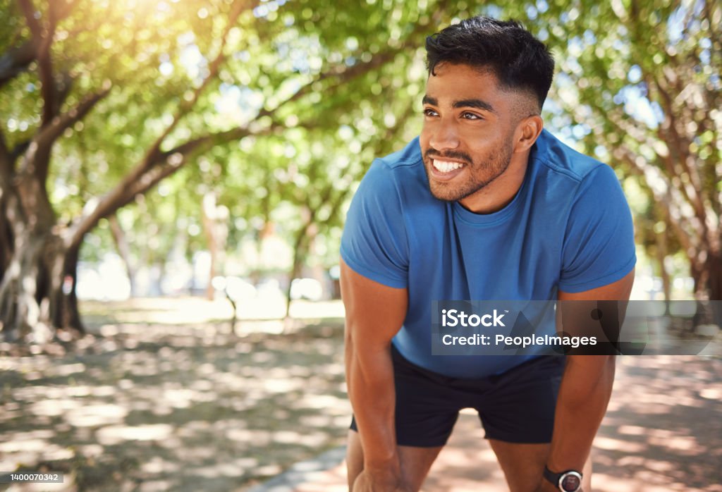 Happy athletic mixed race man smiling while standing with his hands on his knees in a park. Hispanic male resting after his run. Fit man pleased with his performance while exercising outdoors Men Stock Photo