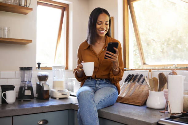 istock Young cheerful mixed race woman drinking a cup of coffee while using her phone alone in the morning in the kitchen. One happy hispanic female  smiling and enjoying a cup of tea while using social media on her phone at home 1400070899