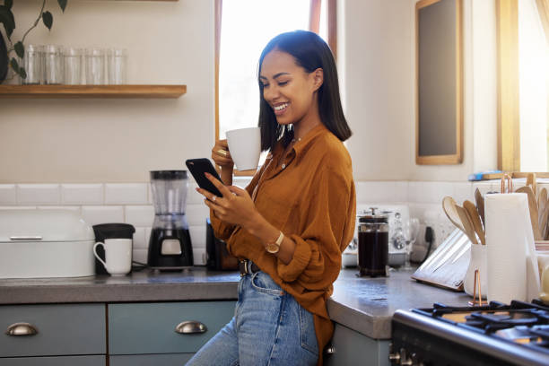 istock Young happy mixed race woman drinking a cup of coffee while using her phone alone in the morning in the kitchen. One hispanic female smiling and enjoying a cup of tea while using social media on her phone at home 1400070908
