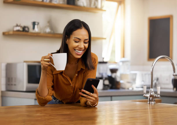 istock Young happy mixed race woman drinking a cup of coffee while using her phone alone in the morning in the kitchen. One cheerful hispanic female smiling and enjoying a cup of tea while using social media on her phone at home 1400071046