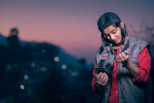 An Asian/Indian confident young woman photographer removes dust from her camera lens through a manual air blower pump in this outdoor dusk image with copy space. She wears a black cap and a warm sleeveless jacket with big pockets. She shoots in the hills of Himachal Pradesh.