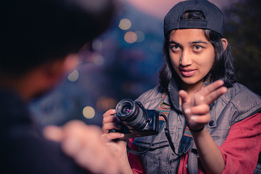 An Asian/Indian confident young woman photographer instructs the male model for the photoshoot In this outdoor dusk image. The photographer is wearing a black cap and a warm sleeveless jacket with big pockets. She shoots in the hills of Himachal Pradesh.