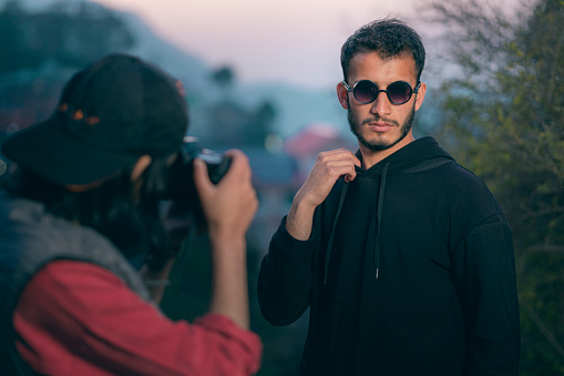 An Asian/Indian confident male model poses for a young woman photographer in this outdoor dusk image. The model wears a hooded sweatshirt and round sunglasses. They shoot in the hills of Himachal Pradesh.