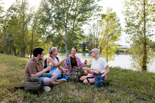 A medium close up of a group of friends sitting and having a picnic whilst on a hike in Toulouse in the South of France. They are at a picturesque riverside area.