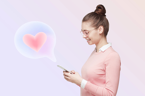 Profile view of cute romantic female with hair knot in pink clothes chatting in dating app for smartphone, heart icon in speech bubble popping from screen, isolated on white background
