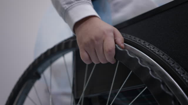 Close-up wheelchair wheel with unrecognizable Caucasian boy hand indoors. Little ill patient sitting on mobility aid device. Health care and disability concept.