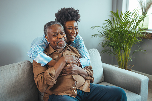 Loving senior man embracing his daughter while sitting on sofa smiling. An attractive young woman embracing her grandfather from back. Senior African American man and granddaughter smiling at the camera