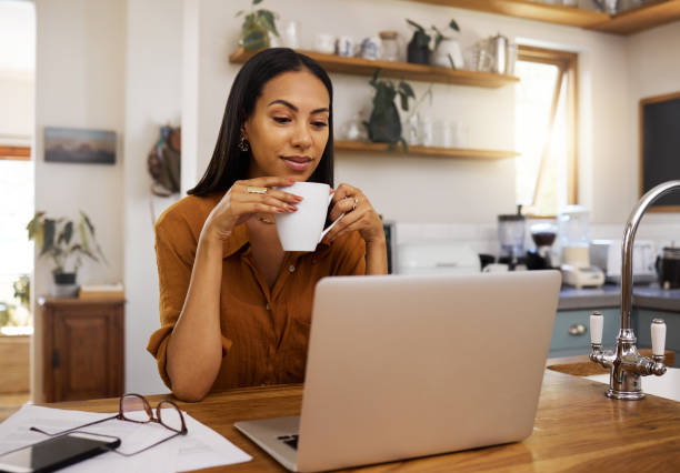 istock Young mixed race woman drinking a cup of coffee while using her laptop alone in the morning in the kitchen. Content hispanic female smiling and enjoying a cup of tea while reading an email on her computer at home 1400080096