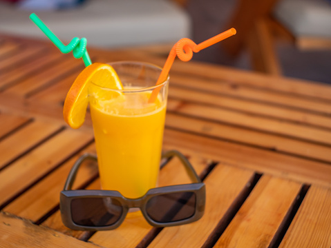 A glass of orange juice with a ripe orange slice and a green straw is on the table and sunglasses are nearby. Copy space.