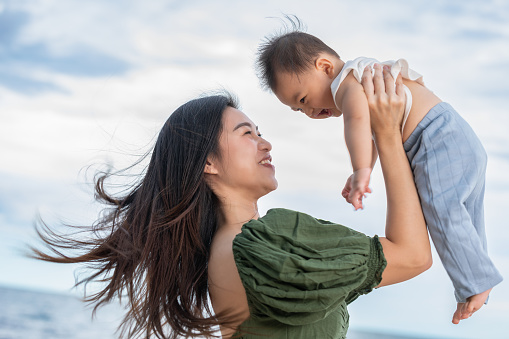 Happy Joyful mother and Son having fun Playing on the Beach together. Asian mom walks by ocean on the beach and throwing up her son in the air at the tropical view. Motherhood Family Concept.