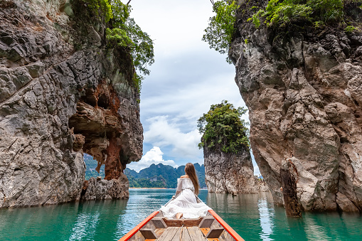 Back View of Young Female Tourist in Dress and Hat at Longtail Boat near Famous Three rocks with Limestone Cliffs at Cheow Lan Lake. Travel Woman Sitting on Boat in Khao Sok National Park in Thailand