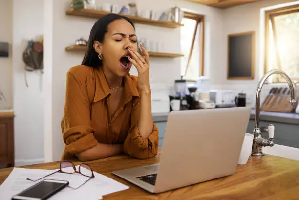 Bored mixed race businesswoman yawning while going through paper and bills and working on a laptop at home. Tired hispanic female businessperson yawning while working from home