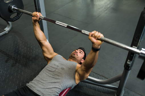 High angle of a sweaty young man lifting heavy weights at the gym and building a muscular body