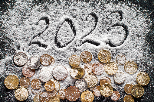 New year concept - close-up of hand written year 2023 and various coins covered with white sea salt such as snow on black background, selective focus. New Year card. The concept of wealth and success