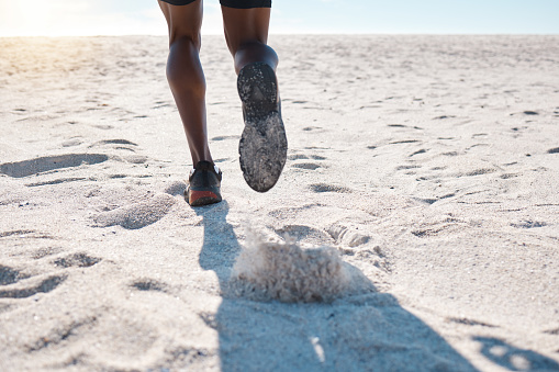 Feet of one black man running and jogging on sand at the beach in the morning for exercise. Closeup of one male athlete from behind doing fast cardio workout with speed to build muscle and endurance