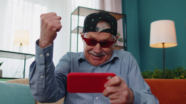 自宅で携帯電話でシューティングゲームのオンラインビデオゲームをプレイして面白い先輩の古い祖父の男を心配して - one senior man only grandfather portrait old ストックフォトと画像