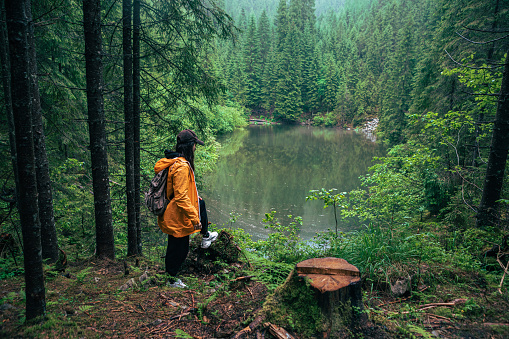 woman hiker with backpack in yellow raincoat looking at mountain lake copy space