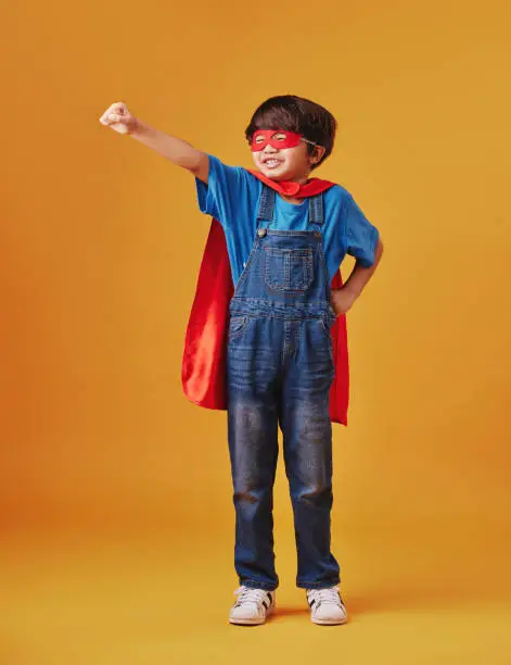 Photo of Adorable little asian boy wearing a mask and a cape while pretending to be a superhero against an orange studio background. Cute happy boy pretending to be a character for halloween