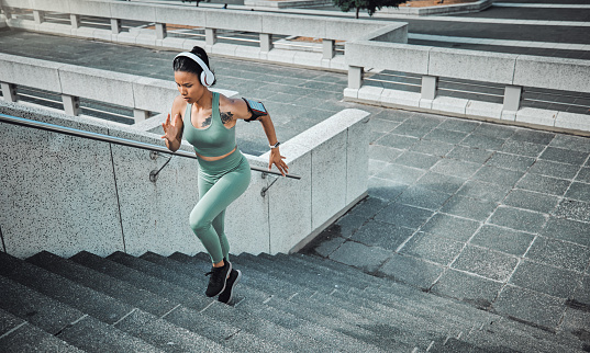 Young mixed race female athlete wearing gymwear and headphones while running up the steps of a building outside. Young sportswoman focused on her speed, body, fitness and cardio health while training