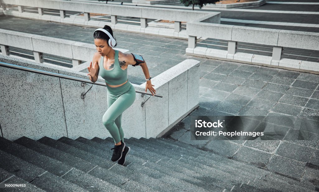 Young mixed race female athlete wearing gymwear and headphones while running up the steps of a building outside. Young sportswoman focused on her speed, body, fitness and cardio health while training Exercising Stock Photo