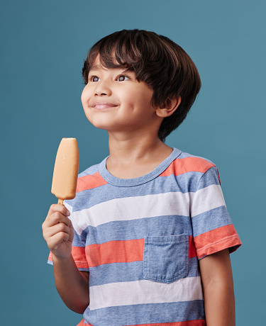 One adorable little asian boy looking happy while enjoying a sweet treat against a blue background. Mixed race child eating a sugar popsicle in summer