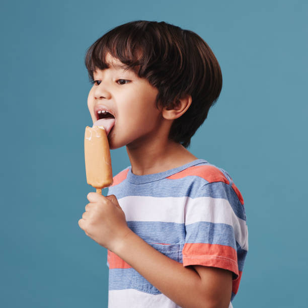 One adorable little asian boy looking happy while enjoying a sweet treat against a blue background. Mixed race child eating a sugar popsicle in summer One adorable little asian boy looking happy while enjoying a sweet treat against a blue background. Mixed race child eating a sugar popsicle in summer licking photos stock pictures, royalty-free photos & images