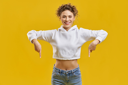 Front view of pretty woman showing down, pointing by fingers. Attractive girl standing, smiling, looking at camera, wearing white khudi and blue jeans. Concept of modern life.