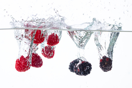 raspberries and blackberries fall into the water on a white background