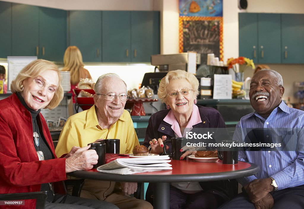 Senior adults drinking coffee in a cafe A group of four seniors sits at a round red table for morning tea from black mugs.  They are all smiling and adorned in colorful clothing.  The kitchen in the background is cream colored with teal cabinetry.  A coffee service station can be seen. Senior Adult Stock Photo