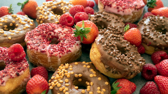 Stock photo showing close-up, elevated view of a display of croissant-doughnuts glazed with chocolate or strawberry fondant icing covered in sprinkles surrounded by fresh strawberries and raspberries, on a blue background.