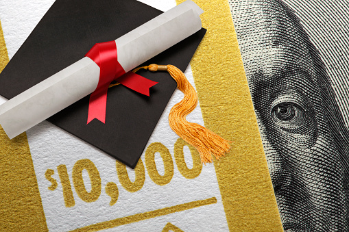 A graduation cap and a rolled up diploma rest on top of a stack of one hundred dollar bills bundled by a $10,000 wrapper.