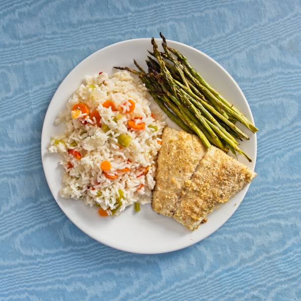 Baked Red Snapper fish plated with seasoned rice and asparagus. Tasty red snapper served with rice and mixed peppers with grilled asparagus. On white plate and blue tablecloth.
