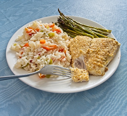 Baked Red Snapper fish plated with seasoned rice and asparagus with fork testing the flaky white fish. Tasty red snapper served with rice and mixed peppers with grilled asparagus. On white plate and blue tablecloth.