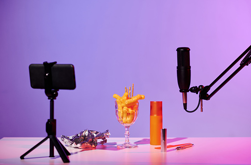 No people still life shot of microphone, smartphone on tripod, glass with crunchy snacks, foil and cosmetic products on table prepared for ASMR