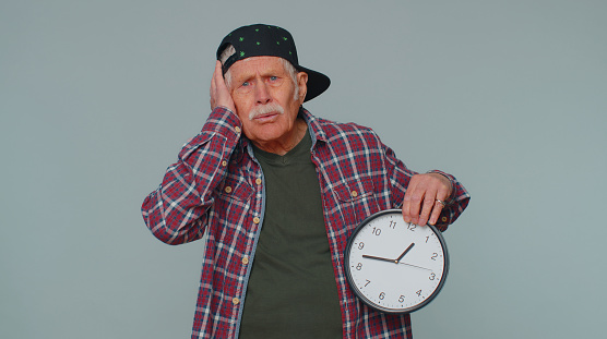 Confused elderly man with anxiety checking time on clock, running late to work, being in delay, deadline. Gray studio background. Serious grandfather looking at hour, minutes, worrying to be punctual