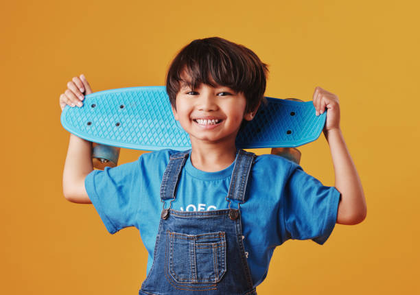 An adorable little asian boy looking happy while holding his skateboard against an orange background. Cute boy wearing casual clothes smiling as he carries his skateboard. Cute child having fun stock photo