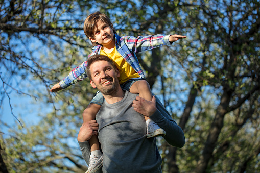 A young boy with outstretched arms sits on the father's shoulders and imagine a flight. Outdoors portrait of cheerful father with son.