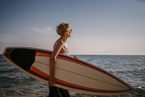 Mature woman surfer walking on the beach by the sea and carrying her surfboard on a sunny day outdoors.