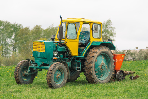 Preili, Latvia - May 14 2022: Plowing and sowing the soil with an old farm tractor Belarus in an agricultural field in spring, with old traditional wooden houses