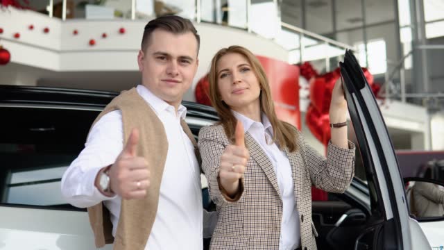 Happy couple at a car dealership with a new car in the background. The guy gave his girlfriend a car for valentine's day