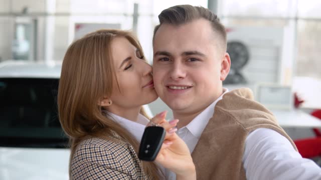 Happy couple taking a selfie at a car dealership with a new car in the background. Man and woman showing keys of his new car in dealership