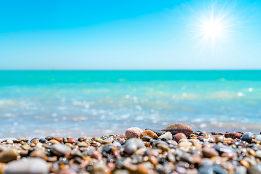 Summer abstract backgrounds: blurred tropical turquoise beach with white waves at background with focused sea pebbles at the bottom of the frame.