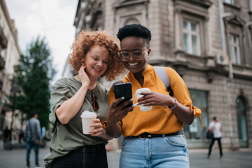Two happy female friends having fun together in the city, having a cup of coffee and using a smart phone to get around
