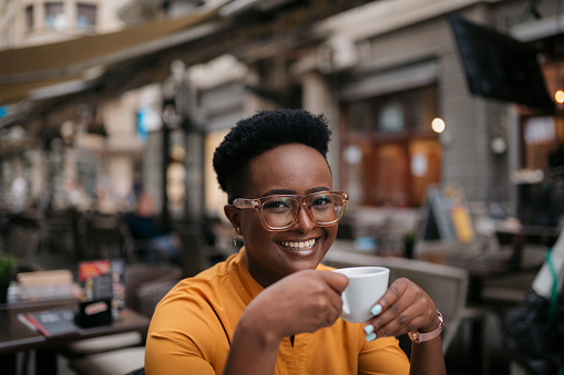 Portrait of a happy young woman sitting in an outdoor coffee shop and enjoying a cup of espresso with a toothy smile on her face while looking at the camera
